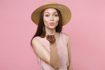Pretty charming beautiful attractive young brunette woman 20s wearing pink summer dotted dress hat posing blowing sending air kiss looking camera isolated on pastel pink background studio portrait.