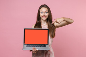 Excited young brunette woman in pink summer dotted dress posing point index finger on laptop pc computer with blank empty screen mock up copy space isolated on pastel pink background studio portrait.