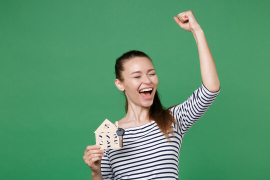 Happy Young Brunette Woman 20s Wearing Striped Casual Clothes Posing Standing Holding House Bunch Of Keys Doing Winner Gesture Keeping Eyes Closed Isolated On Green Color Background, Studio Portrait.