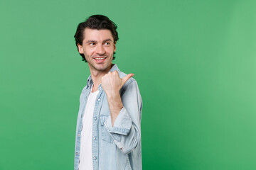 Side view of smiling young brunet man 20s wearing casual clothes white t-shirt denim shirt posing stand pointing thumb aside on mock up copy space isolated on green color background studio portrait.