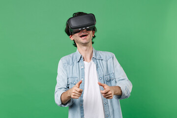 Cheerful young brunet man 20s wearing casual clothes white t-shirt denim shirt posing standing watching in headset play game with joystick isolated on green color wall background studio portrait.