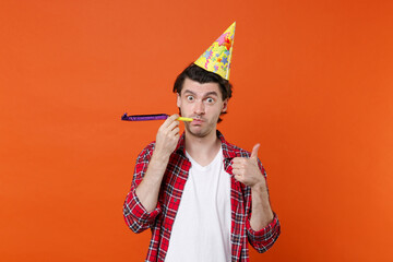 Funny young brunet man 20s in casual clothes white t-shirt red checkered shirt birthday hat posing blowing in pipe showing thumb up looking camera isolated on orange color background studio portrait.