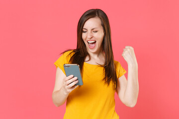 Happy joyful young brunette woman 20s wearing yellow casual t-shirt posing using mobile cell phone typing sms message doing winner gesture isolated on pink color wall background studio portrait.