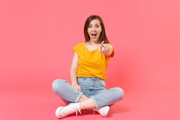 Full length portrait of cheerful funny excited beautiful young brunette woman 20s wearing yellow t-shirt sitting on floor pointing index finger on camera isolated on pink color wall background studio.