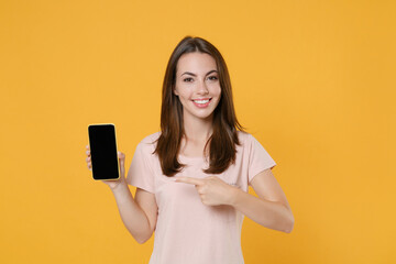 Naklejka premium Smiling young brunette woman in pastel pink casual t-shirt posing pointing index finger on mobile cell phone with blank empty screen mock up copy space isolated on yellow background studio portrait.