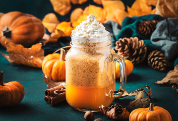 Pumpkin spiced latte or coffee in glass jar on blue table. Autumn or winter hot drink in festive natural table setting with orange leaves, spices, small pumkins, pine cones