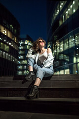 A girl in a white t-shirt and jeans is sitting on the stairs. Portrait of a girl in the style of the 90s. A woman with curls and pink lipstick on the background of an urban landscape.