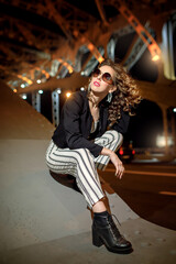 A curly-haired girl wearing sunglasses and white earrings is sitting on a bridge support. Portrait of a young woman in the style of the 90s. Night portrait of a model on the bridge.