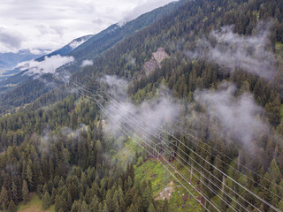 Aerial view of power line through mountain valley in Switzerland. Fog and mist hanging in the air. Cloudy conditions. Concept of power transmission.