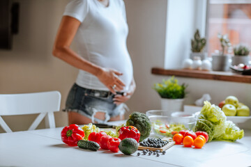 Young woman's pregnant body posing near the kitchen table with vegetables.