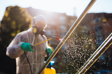 Frontline male worker with face mask and protective workwear using pressure sprayer to disinfect children's playground. Virus pandemic protection and prevention concept.