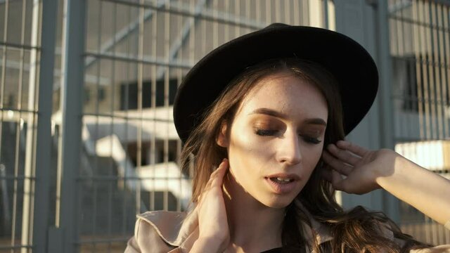 Beautiful girl model stands and puts on a hat on the street. Brunette with long hair in a black hat and brown cloak. Portrait frame from the back