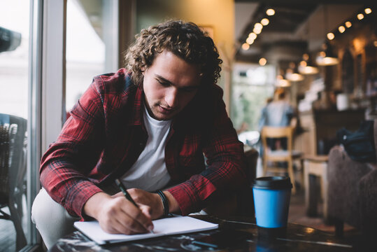 Serious caucasian male student 20s sitting in coffee shop learning and doing homework task, pensive hipster guy in casual outfit writing article and publication in blog i notepad in cafe interior