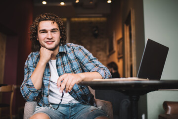 Half length portrait of casually dressed male student sitting in cafe interior with technology for learning, prosperous 20s hipster guy freelancer satisfied with remote job looking at camera