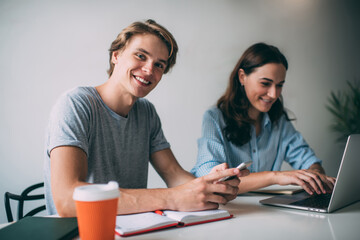 Portrait of cheerful caucasian man holding mobile phone chatting while his female colleague make research on laptop computer for homework in college, millennial hipster guys enjoying study via gadge.