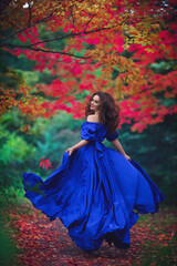 Romantic portrait of a beautiful young girl in a long ultramarine blue dress running away on the background of an autumn рark with red leaves