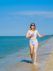 Fototapeta premium A young girl runs along the sandy sea beach along the surf. Splashes of sea water. The girl is wearing a white swimsuit and sunglasses. Freedom and carelessness. Outdoor fitness. Sunny day. Copy space