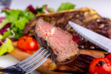 A slice of steak impaled on a fork on the background of a plate of meat.