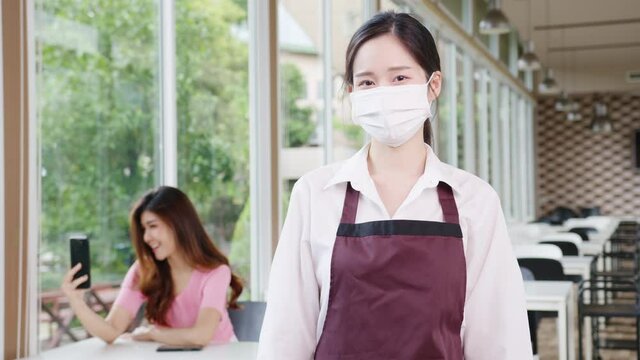 Young Asia female restaurant staff wearing protective face mask holding alcohol and wet wipe before welcome customer with customer in background. Lifestyle new normal restaurant after corona virus.