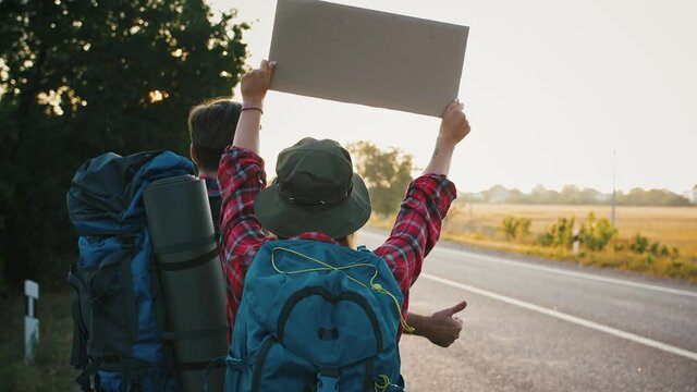 Couple of tourists unsuccessfully trying to catch car, standing with poster on road, back view