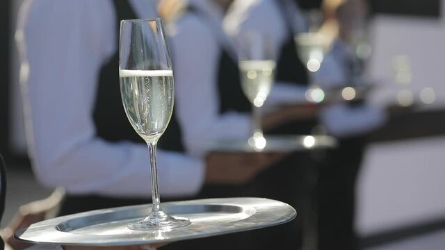 The waiter holds a tray with a glass of champagne in his hand, small bubbles of gas are released in the champagne and are illuminated by the sun. 
