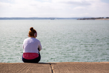 A teenager sitting alone on a wall listening to music on headphones looking at a view of the sea