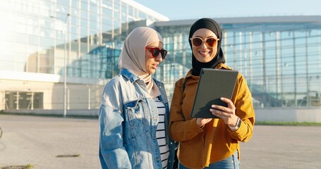 Arabian pretty females in headscarves standing outdoors at aeroport, watching something on tablet...