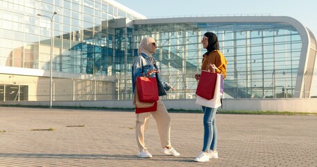 Two muslim stylish women in headscarves standing outdoors at aeroport, talking and holding shopping packets. Stylish females at street chatting. Shoppers. Best friends having conversation.