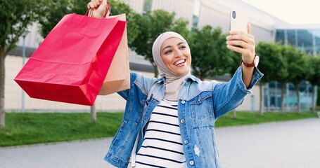 Pretty muslim happy woman in traditional headscarf holding bags after shopping and showing to smartphone camera while videochatting. Outdoors. Beautiful cheerful girl in hijab talking in videochat.