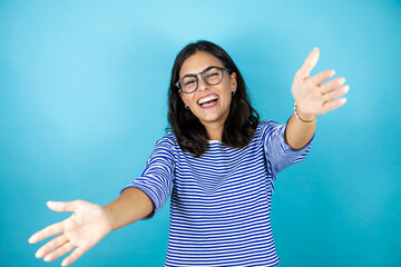 Pretty woman wearing glasses standing over insolated blue background looking at the camera smiling with open arms for hug.