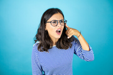 Pretty woman wearing glasses standing over insolated blue background doing phone gesture with hand and fingers like talking on the telephone