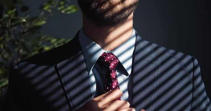 Close up of mature bearded man adjusting trendy tie while standing at modern office. Concept of successful career, business and finance.