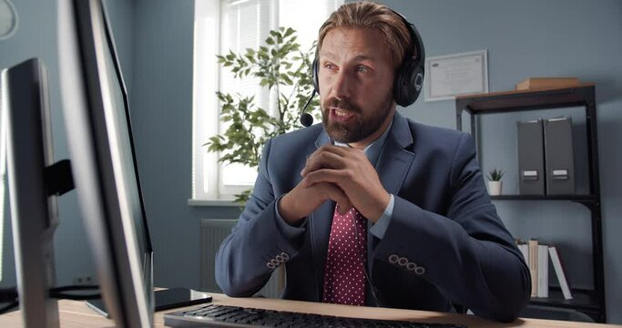Confident mature man in headset having video conference on computer while working at office. Bearded businessman in suit talking with partners online.