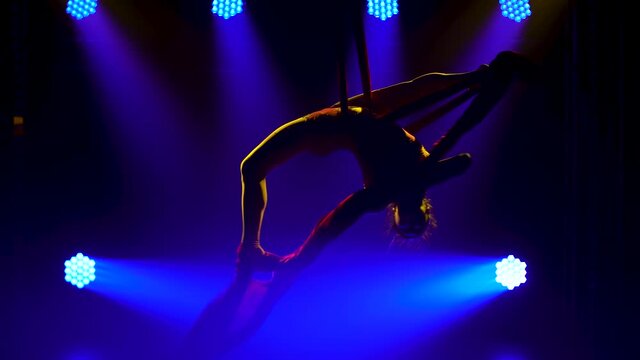 Young woman doing show acrobatic trick on air silk. Exciting acrobatic show in a dark studio with blue stage lighting. Silhouette of a slim body. Close up.