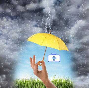 Insurance Concept. Man Covering Medical Kit Illustration With Yellow Umbrella During Storm, Closeup