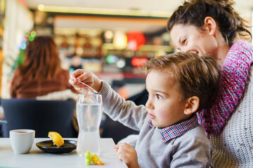 Side view on single mother and son sitting in cafe small boy holding plastic spoon playing with his drink in glass - growing up family concept