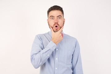 European man dressed in formal shirt and bow tie poses against white studio background Looking fascinated with disbelief, surprise and amazed expression with hands on chin