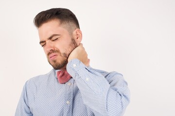 Fototapeta premium European man dressed in formal shirt and bow tie poses against white background Suffering of neck ache injury, touching neck with hand, muscular pain