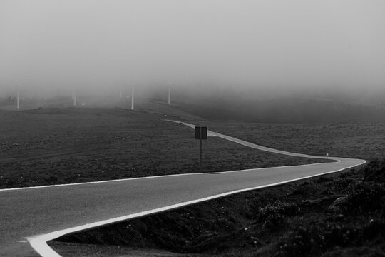 Dramatic black and white shot of a solitary road covered by the mist in the mountains with windmills at the end of it