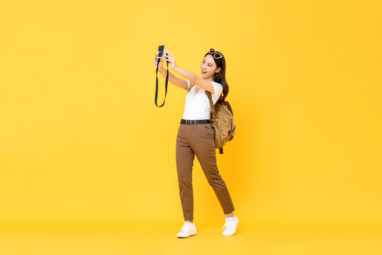 Young pretty Asian tourist girl taking selfie with digital camera in yellow isolated background