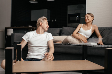 a man in casual clothes fixing a table in his living room and talking to his girlfriend, who sits on the sofa and watches. Family everyday life. Young married couple.