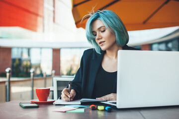 Caucasian entrepreneur with blue hair working in a cafeteria at the laptop and making some notes in a book while drink a coffee