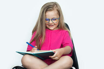 Blonde caucasian girl with eyeglasses writing something in a book while wear red shirt on a white studio wall and sitting in armchair