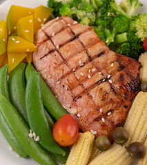 Chicken steak topped with white sesame, peas, tomatoes, broccoli and pumpkin in a white plate.