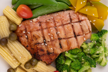 Chicken steak topped with white sesame, peas, tomatoes, broccoli and pumpkin in a white plate.