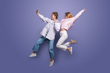 Two charming women in bright clothes and blonde hair jumping on a violet studio wall and making a selfie using a phone
