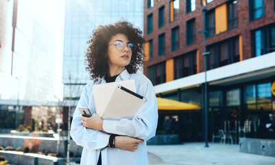 Front view photo of a modern it woman with curly hair and eyeglasses posing in the city with a...