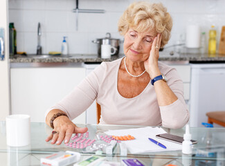 Confused senior woman sitting at kitchen table among a lot of medicines