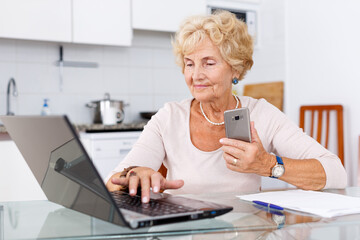 Attractive mature woman sitting at kitchen table and using laptop and smartphone