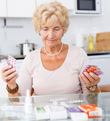 Positive senior woman sitting at kitchen table among a lot of medicines